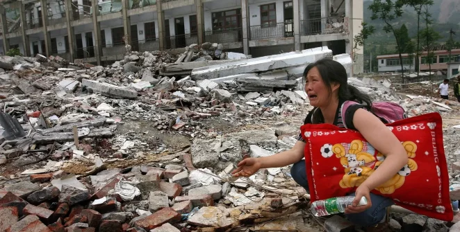 A distraught woman kneels among the ruins of a building destroyed by an earthquake in China, clutching a red pillow with a cartoon, her face expressing shock and sorrow.