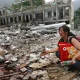 A distraught woman kneels among the ruins of a building destroyed by an earthquake in China, clutching a red pillow with a cartoon, her face expressing shock and sorrow.
