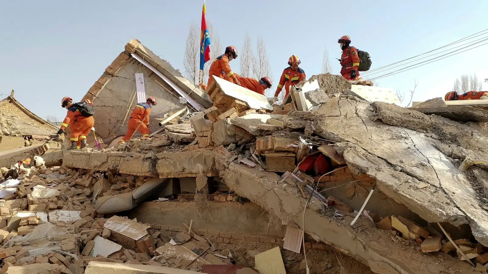 Rescue workers in orange uniforms search through the wreckage of a building brought down by an earthquake in China, with a national flag visible in the background, symbolizing a coordinated disaster response effort.