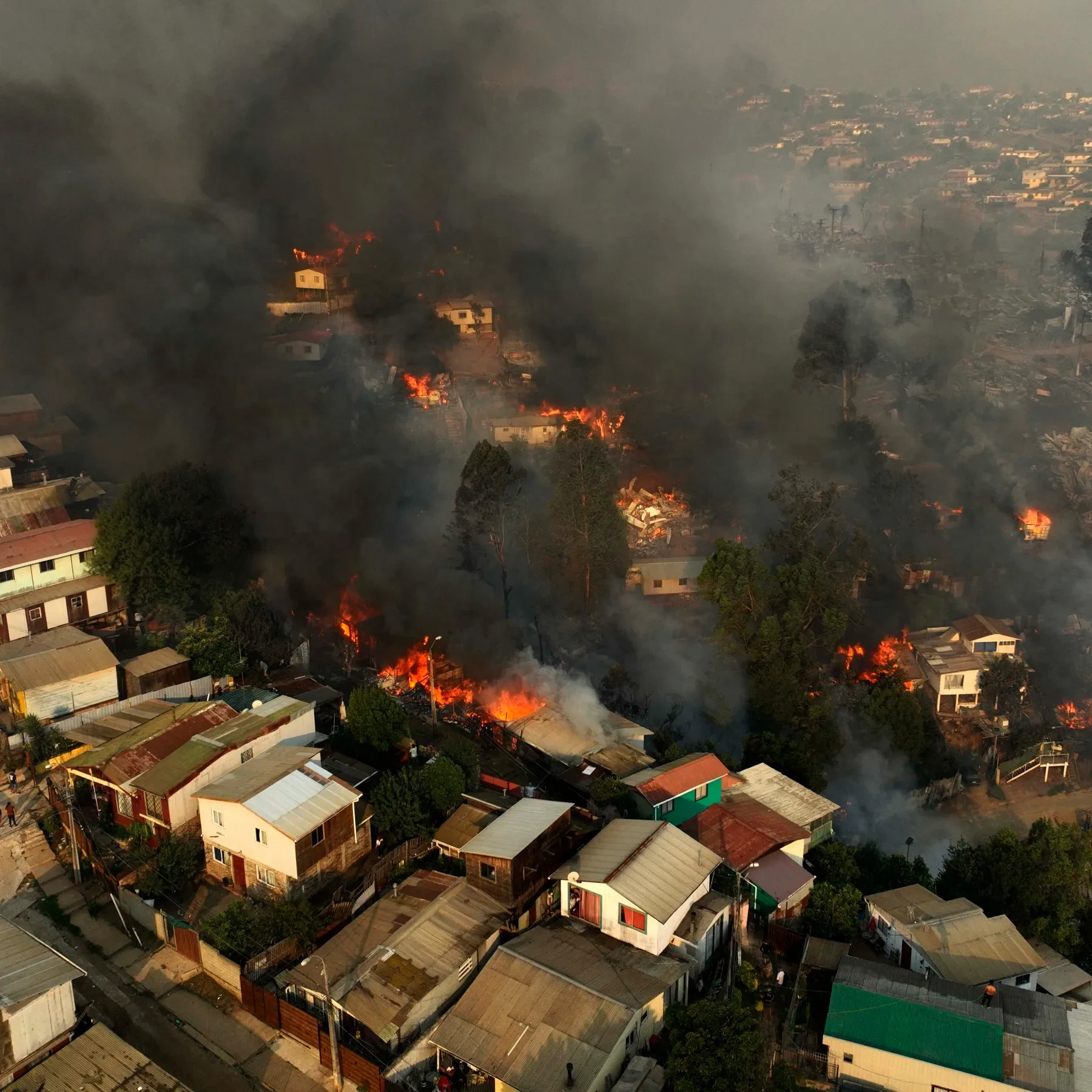 Aerial view of a Chilean neighborhood engulfed in flames from a severe wildfire, with thick smoke rising above the burning houses and trees.