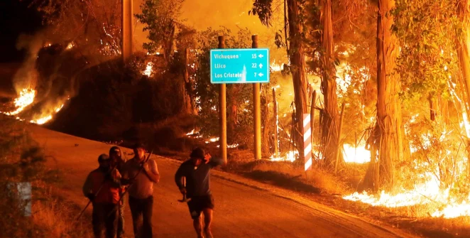 A group of people hastily evacuate on foot at night as a wildfire rages beside the road, illuminating a signpost with directions to nearby towns.