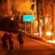A group of people hastily evacuate on foot at night as a wildfire rages beside the road, illuminating a signpost with directions to nearby towns.